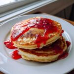 Two fluffy pancakes drizzled generously with bright red guava syrup, served on a white plate.