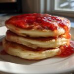 A stack of three fluffy pancakes generously drizzled with bright red guava syrup.