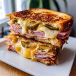 Close-up of a halved Sizzling Cuban Grilled Cheese sandwich stacked high with ham, melted cheese, and pickles.