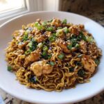 A close-up of a white bowl filled with Sticky Garlic Chicken Noodles, topped with sesame seeds and chopped green onions.