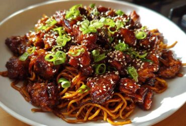 Close-up of Sticky Garlic Chicken Noodles coated in a dark glaze, topped with sesame seeds and green onions.