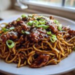 A close-up of Sticky Garlic Chicken Noodles coated in dark sauce, topped with sesame seeds and green onions.