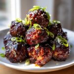 A close-up of a pile of Sticky Hoisin Meatballs Appetizer, glazed dark brown and topped with sesame seeds and sliced green onions.