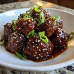 A close-up of Sticky Hoisin Meatballs Asian Appetizer, coated in a dark, glossy sauce and sprinkled with sesame seeds and green onions.