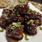 A stack of glossy Sticky Hoisin Meatballs Asian Appetizer coated in dark sauce, garnished with sesame seeds and green onions.