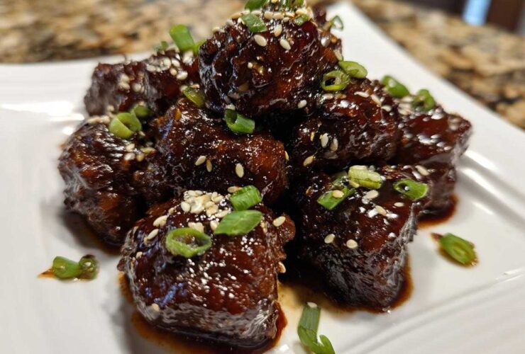 A stack of glossy Sticky Hoisin Meatballs Asian Appetizer coated in dark sauce, garnished with sesame seeds and green onions.