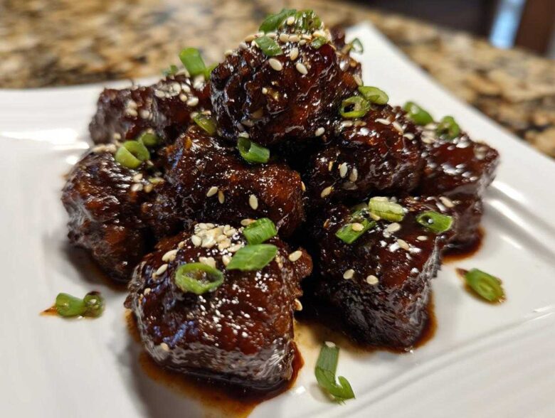 A stack of glossy Sticky Hoisin Meatballs Asian Appetizer coated in dark sauce, garnished with sesame seeds and green onions.