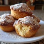 A stack of four golden Vanilla Chai Muffins dusted heavily with powdered sugar on a white plate.