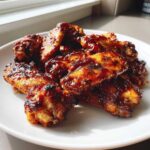 A close-up of several sticky, caramelized BBQ Chicken Wings piled on a white plate, glistening under natural light.