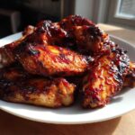 A close-up of sticky, caramelized BBQ Chicken Wings piled high on a white plate, glistening under natural light.