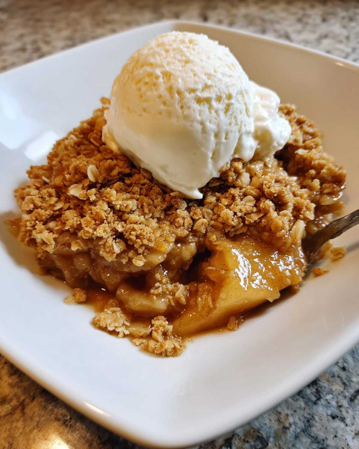 A close-up of a warm serving of Apple Crisp topped with a scoop of melting vanilla ice cream in a white bowl.