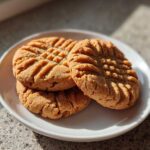 Three golden brown Peanut Butter Cookies stacked slightly on a white plate, showing classic crosshatch fork marks.
