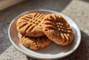 Three golden brown Peanut Butter Cookies stacked slightly on a white plate, showing classic crosshatch fork marks.