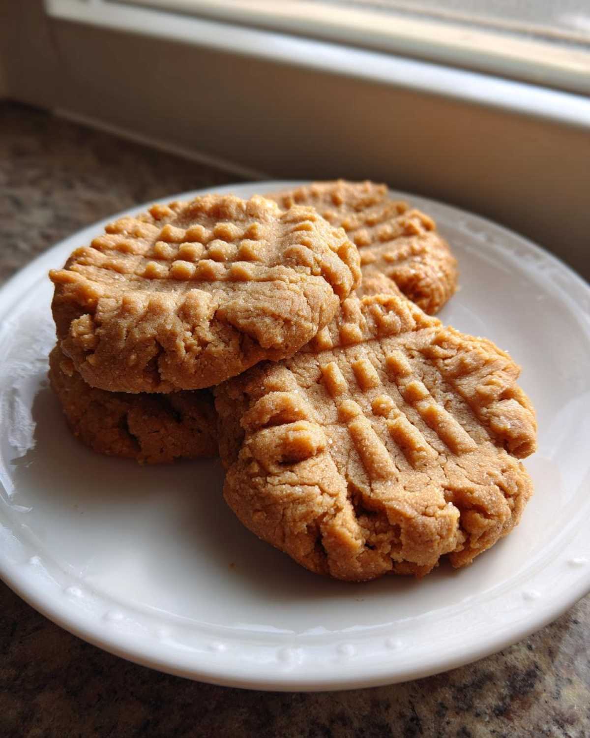 A stack of three classic, fork-pressed Peanut Butter Cookies resting on a white plate near a window.