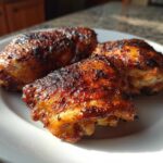 Three crispy, well-seasoned Oven-Baked Chicken Thighs resting on a white plate under bright light.