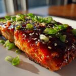 A close-up of a perfectly cooked Gochujang Honey Glazed Salmon fillet topped with sesame seeds and fresh green onions.
