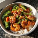 A close-up of a bowl filled with white rice topped with glazed Honey Garlic Shrimp, steamed broccoli, carrots, and green onions.