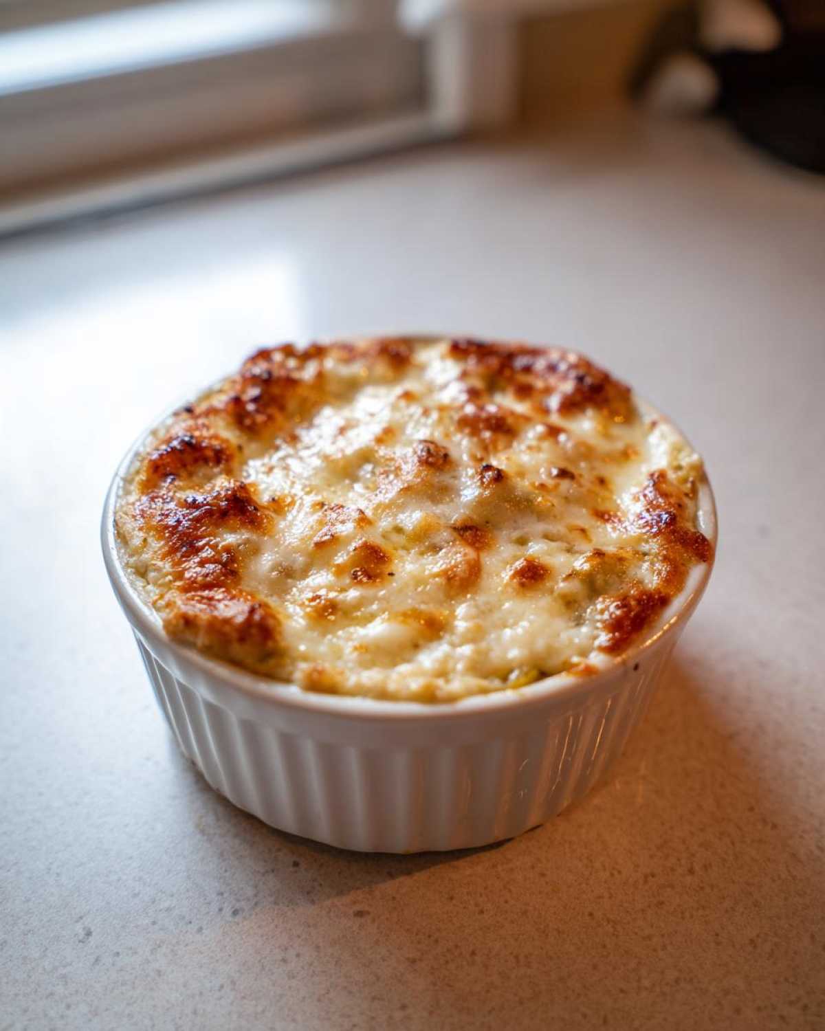 A small, individual serving of hot Baked Pickle Dip topped with bubbly, browned cheese.