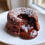 Close-up of a freshly baked Molten Chocolate Lava Cake with a rich, gooey center oozing onto a white plate, dusted with powdered sugar.