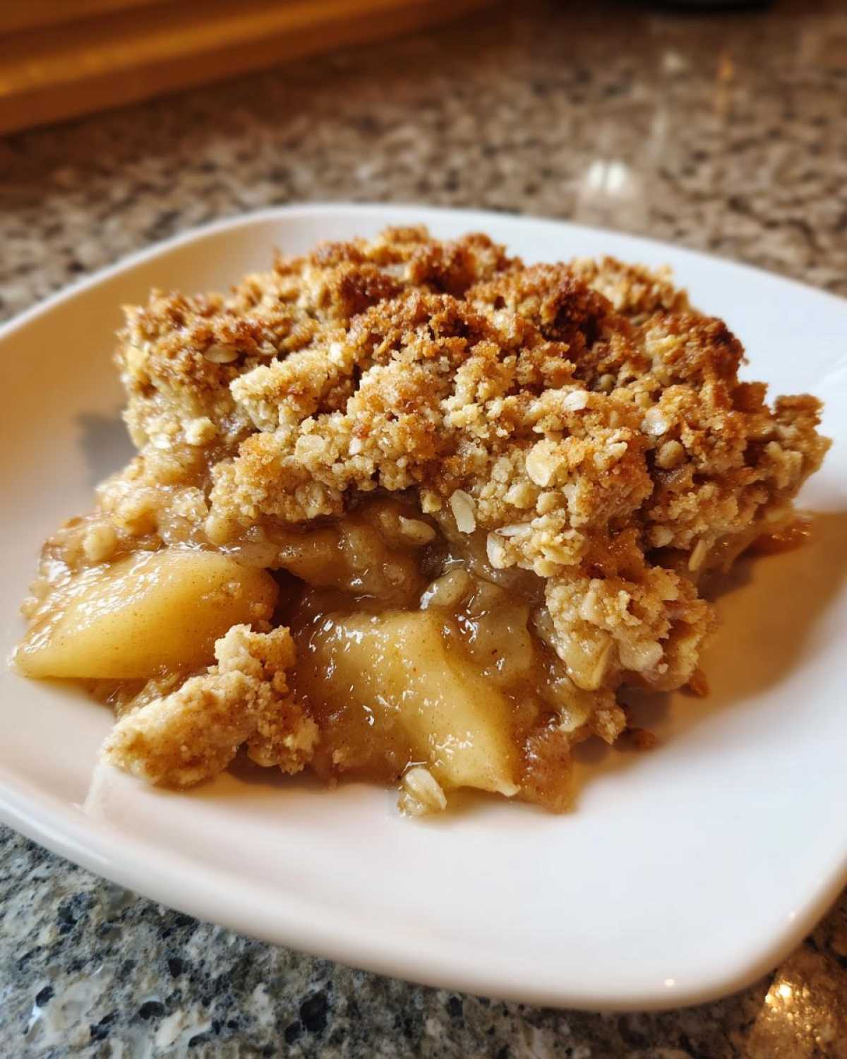 A close-up of a square serving of warm Apple Crisp showing soft, spiced apples beneath a thick, golden, oat crumble topping.
