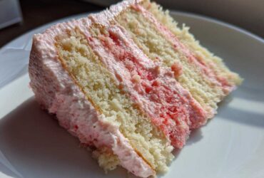Close-up of a moist slice of Strawberry Vanilla Cake with Frosting, showing layers of vanilla cake and pink strawberry filling.