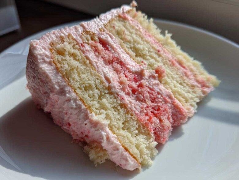 Close-up of a moist slice of Strawberry Vanilla Cake with Frosting, showing layers of vanilla cake and pink strawberry filling.